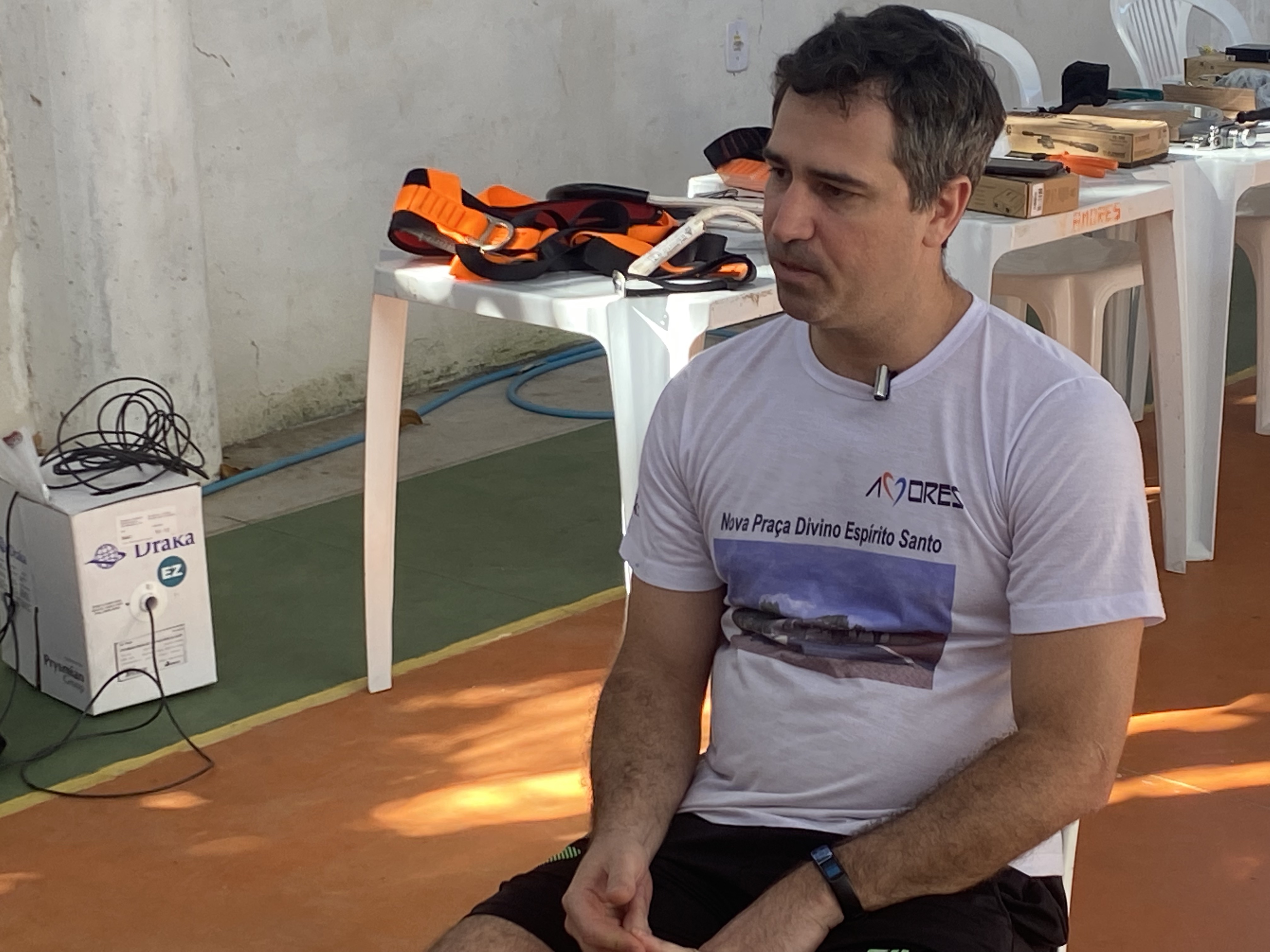 A man in a white t-shirt with a photo of a local square stamped on it, sits on a plastic white chair at the corner of a small public sports court.