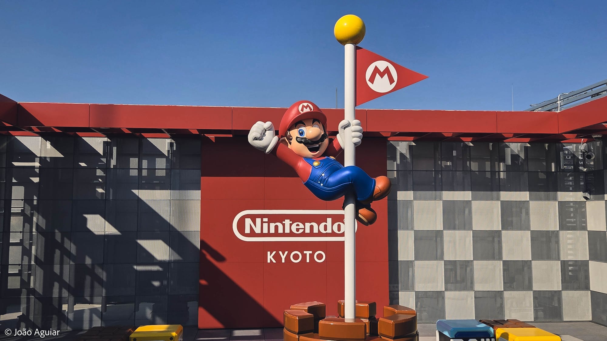 Mario descendo o poste com uma bandeirinha vermelha onde se vê um M. Ao fundo, o logo da Nintendo sobre a palavra Kyoto.