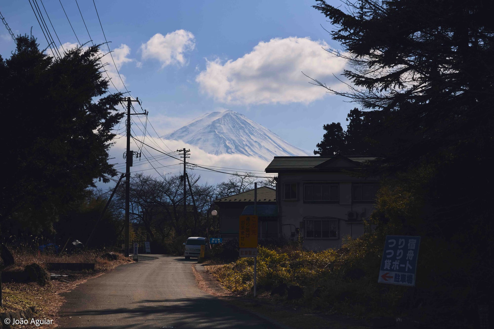Rua pequena carcada por vegetação. À direita, casas sob a sombra de árvores. Ao fundo, o vulcão triangular coberto de neve, o Monte Fuji, chama a atenção.