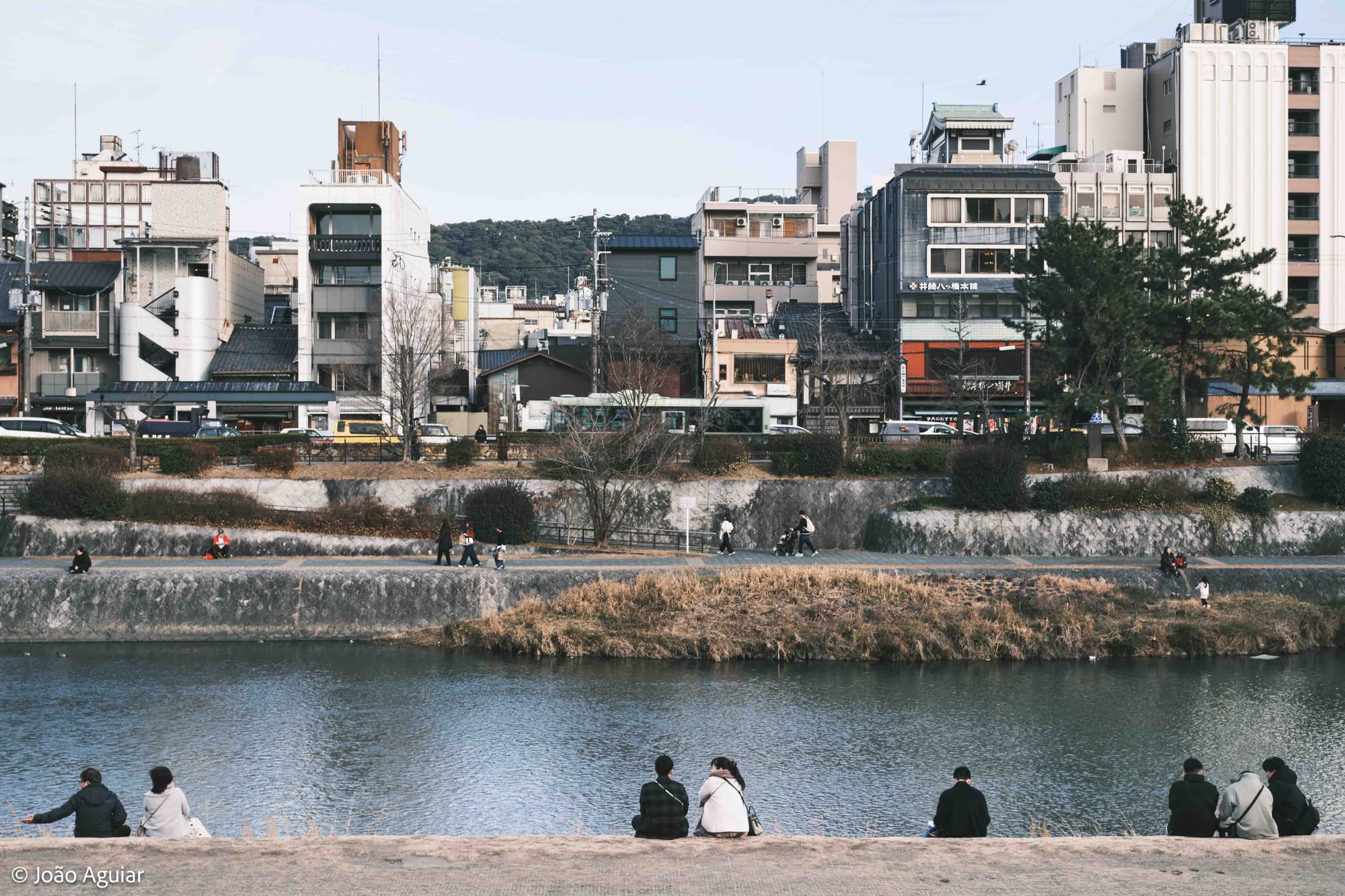 Em primeiro plano, pessoas de costas para a camera sentadas na orla de um rio que se vê diante delas. Ao fundo, prédios pequenos lado a lado.