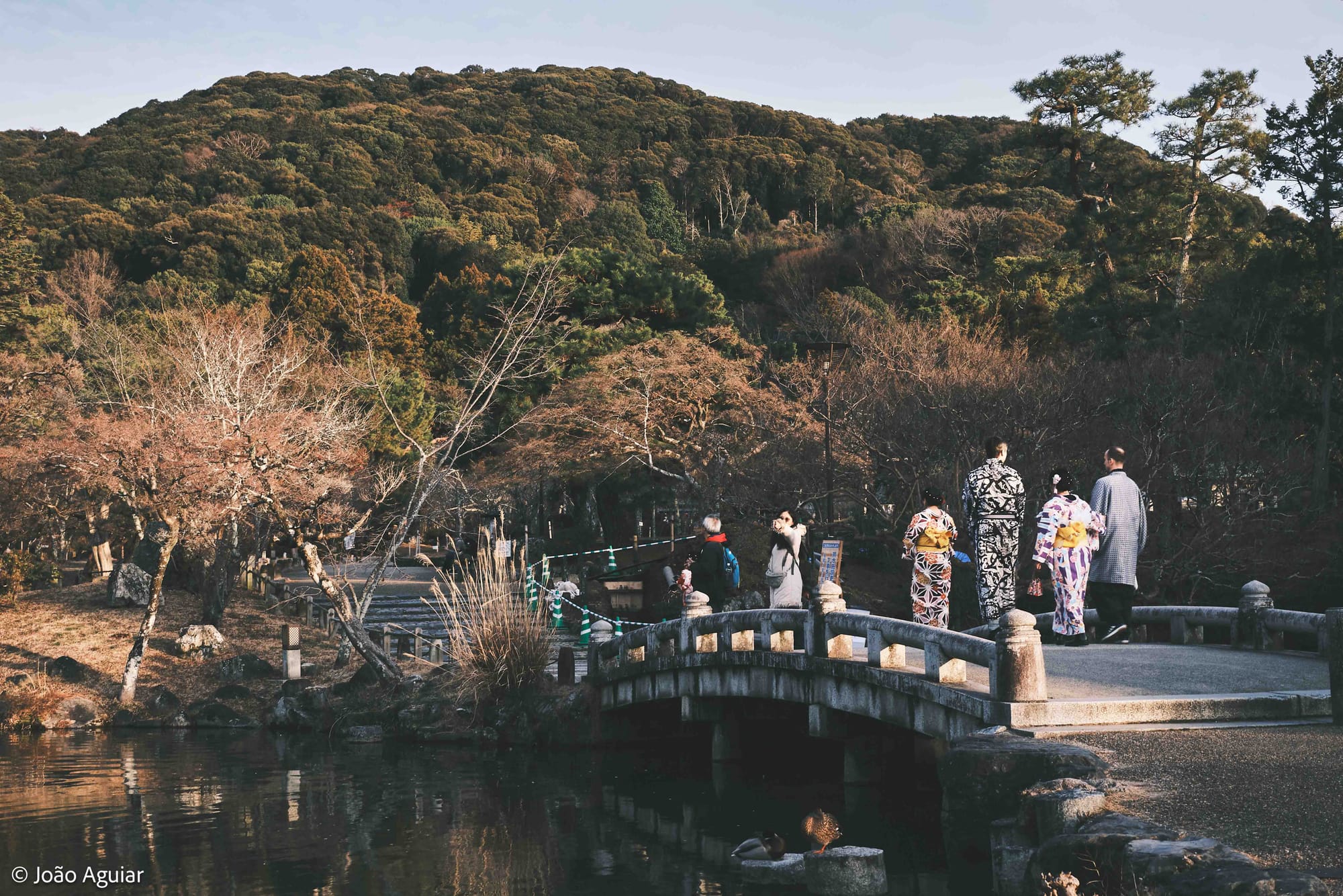 Turistas vestindo quimonos atravessam uma baixa ponte sobre um lago.