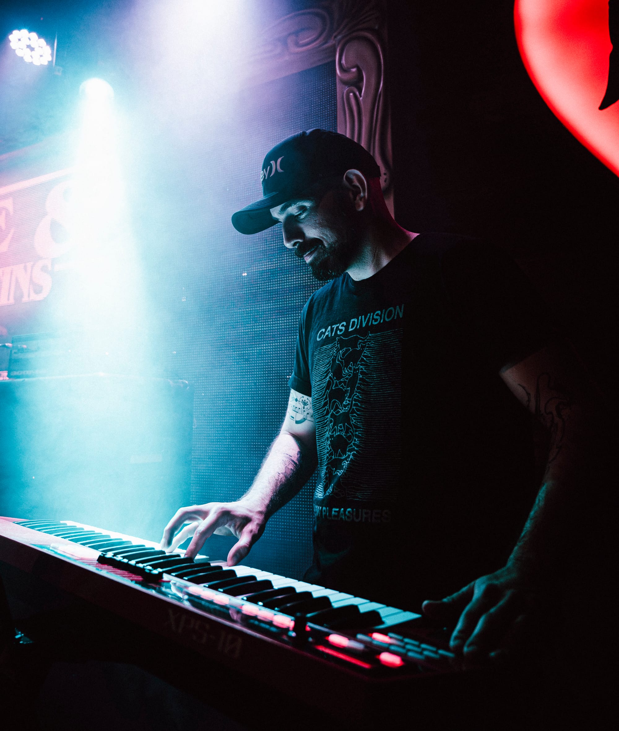 Homem com boné e camiseta preta toca teclado em palco sob forte iluminação azul e branca.