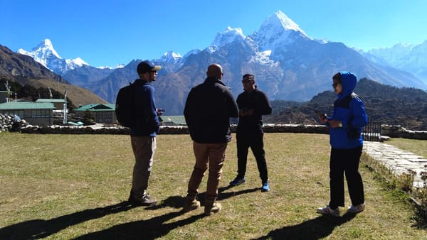 Quatro pessoas conversam de pé sobre um gramado. Elas estão contra a luz, assim que são quase silhuetas. Ao fundo se vê uma g