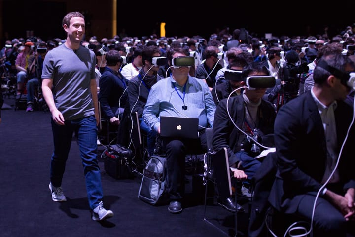 Mark Zuckerberg caminha sorrindo ao lado de uma platéia cheia com os olhos tapados por óculos de realidade virtual.