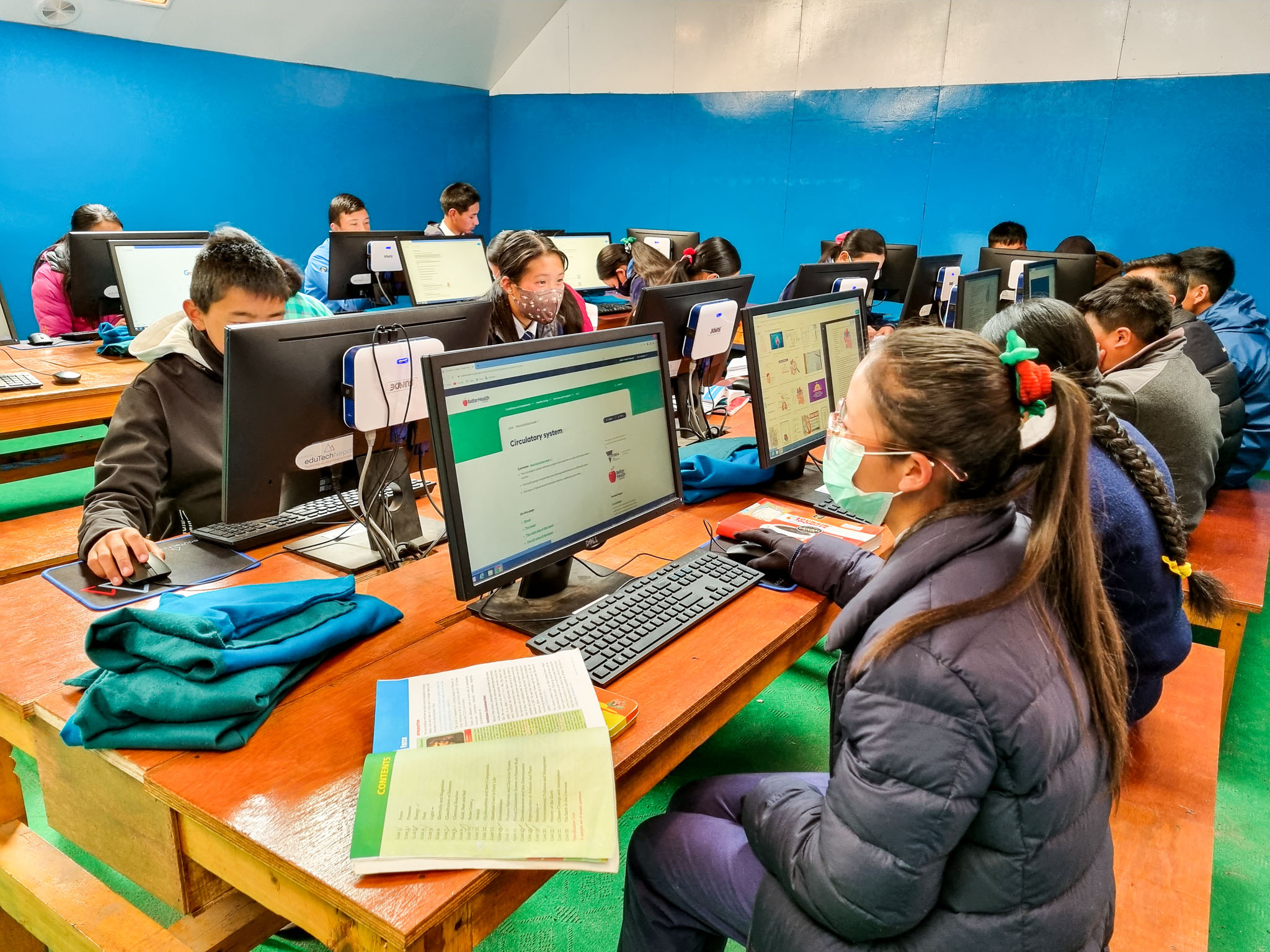 Imagem aberta de um laboratório de informática onde crianças de diferentes idades estão sentadas cada uma em frente a um computador fazendo pesquisas na Internet.