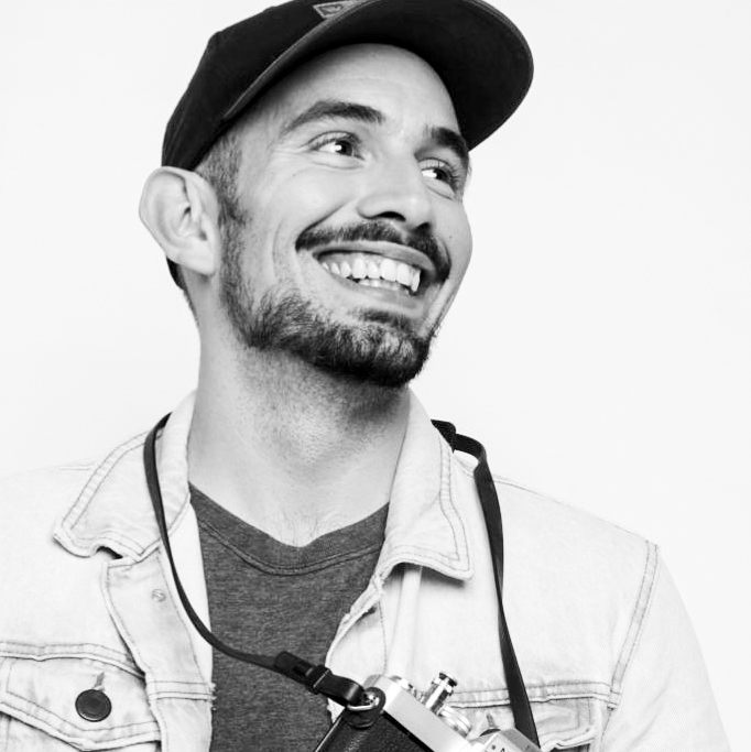 Foto quadrada do jornalista João Aguiar sorrindo. Ele é um homem branco jovem, com barba curta e bigode, usando um boné escuro, uma jaqueta jeans, e uma câmera fotográfica pendurada no pescoço.