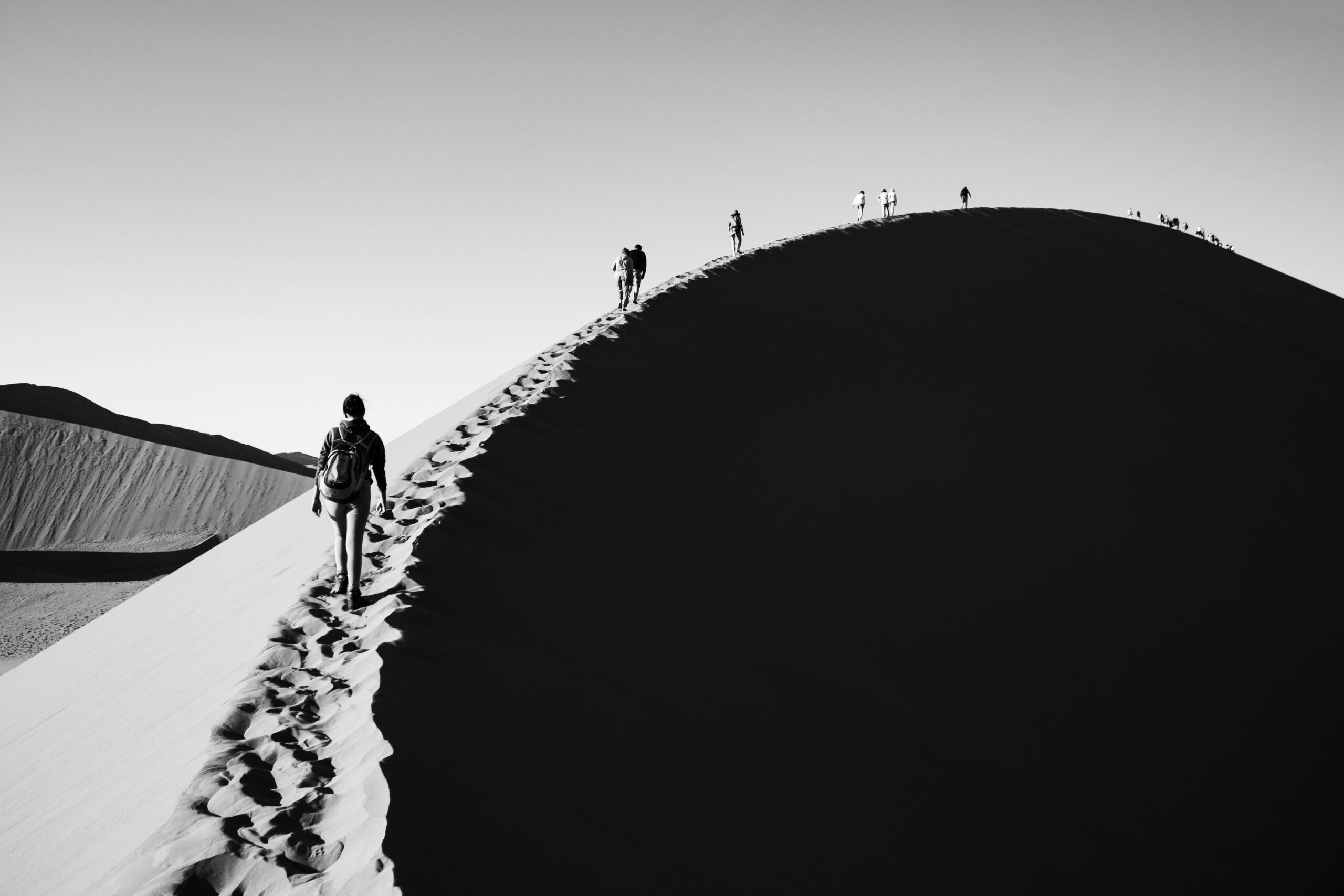 Foto monocromática de pessoas caminhando sobre uma linha de areia no topo de uma duna. O lado esquerdo está iluminado pela luz do sol, enquanto o lado direito tem uma forte sombra preta.