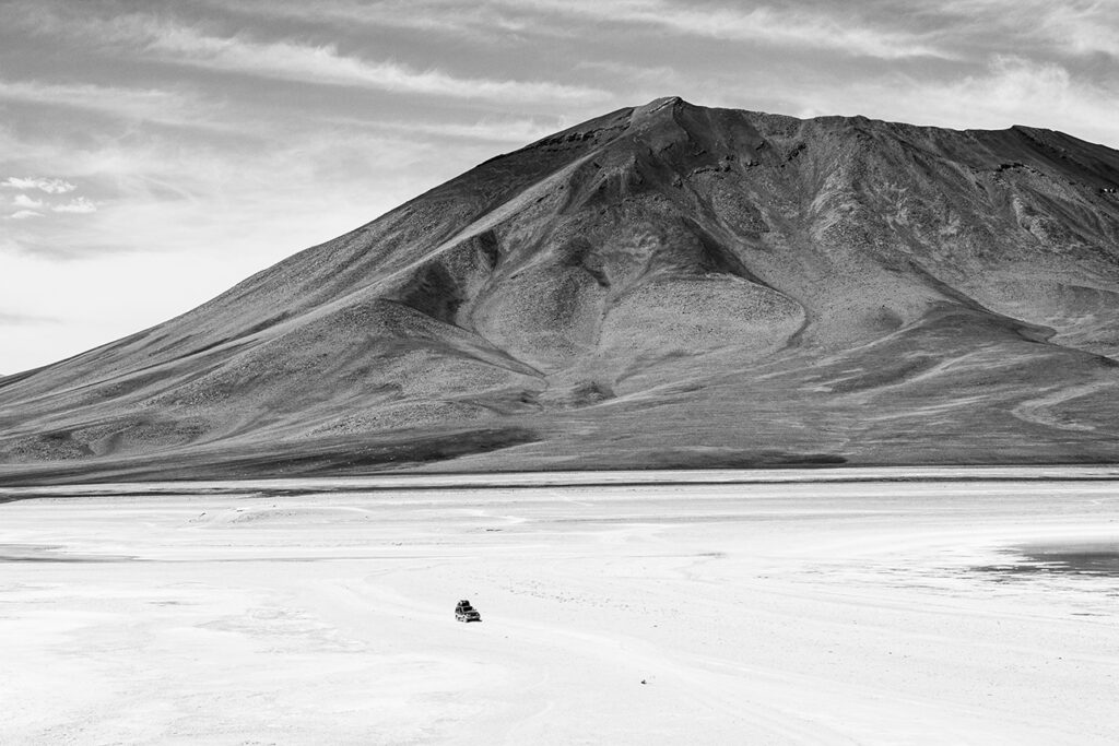 Foto monocromática de um deserto plano com uma grande montanha ao fundo. No primeiro plano, um jeep muito pequeno pode ser visto em meio ao descampado claro. Ao fundo, a montanha está mais escura e em contraste com o tamanho do jeep mostra sua imponência.