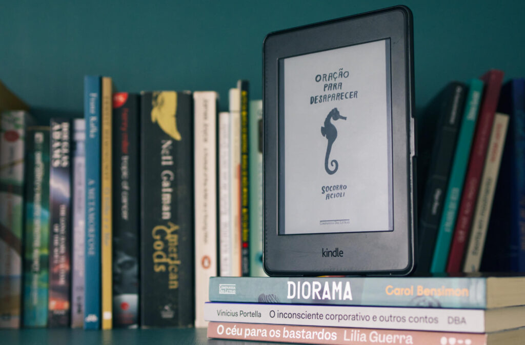 Imagem de um Kindle exibindo a capa do livro "Oração Para Desaparecer" de Socorro Acioli, sobre os livros Diorama, de Carol Bensimon; O Inconsciente Corporativo e Outros Contos, de Vinicius Portella e; O Céu Para Bastardos, de Lilia Guerra. Atrás estão diversos outros livros desfocados.